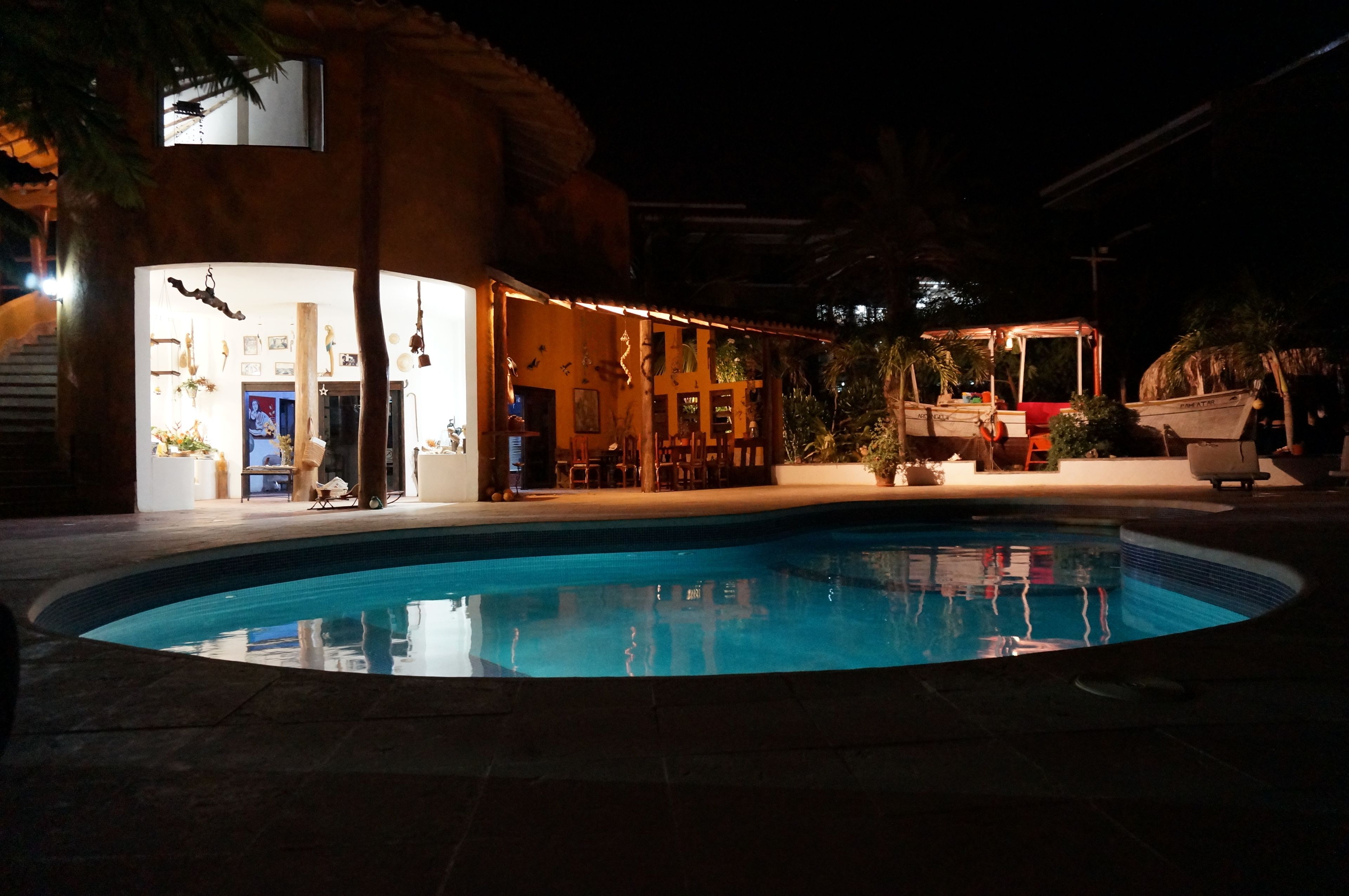 A swimming pool area with a covered seating area and a building with visible interior lighting.