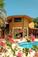 Um edifício de dois andares com piscina, cercado por palmeiras e flores cor-de-rosa.