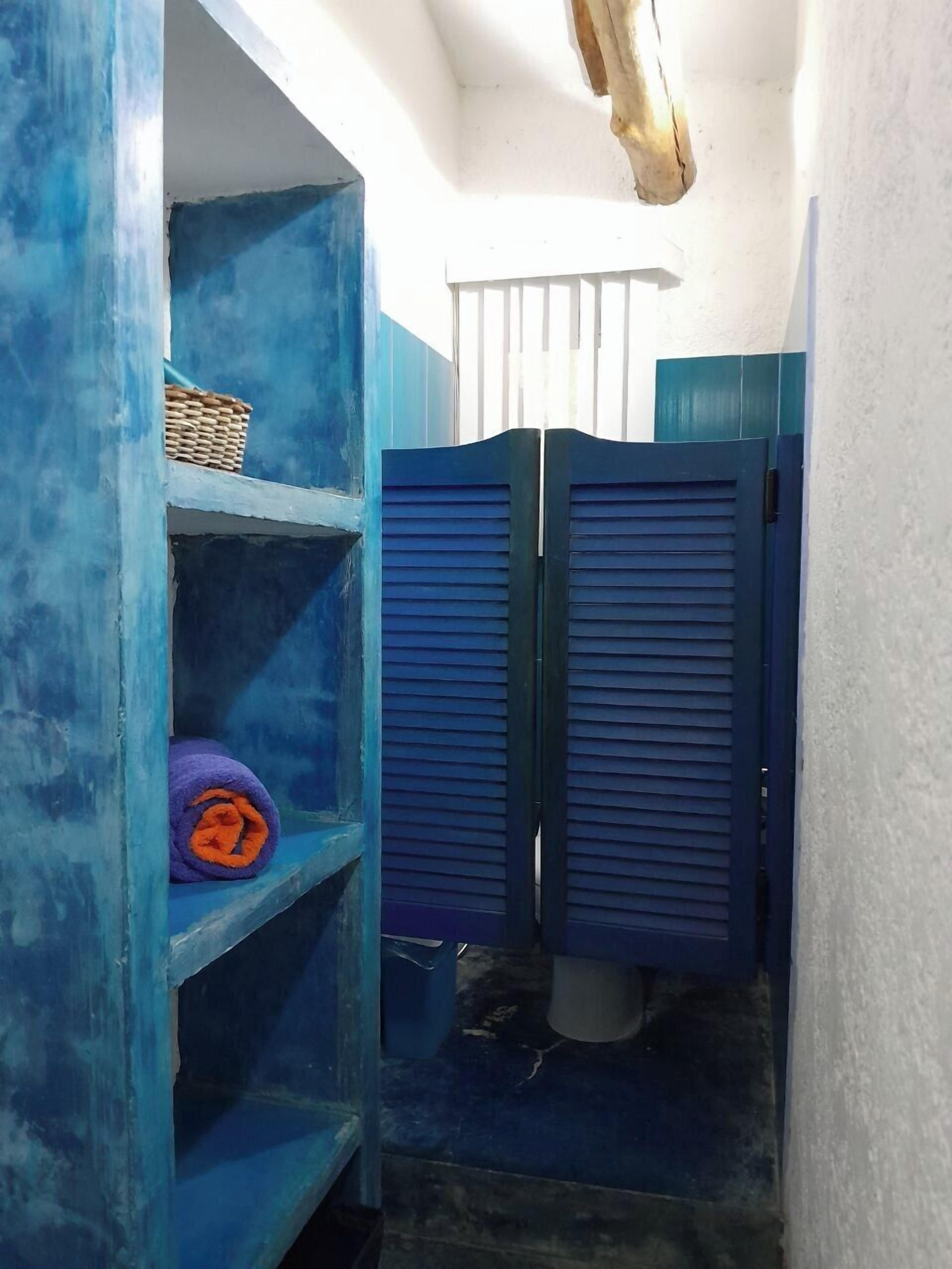 A narrow hallway with blue-painted walls and a white ceiling. There are two blue doors, a basket on a shelf, and a rolled-up purple towel on the floor.
