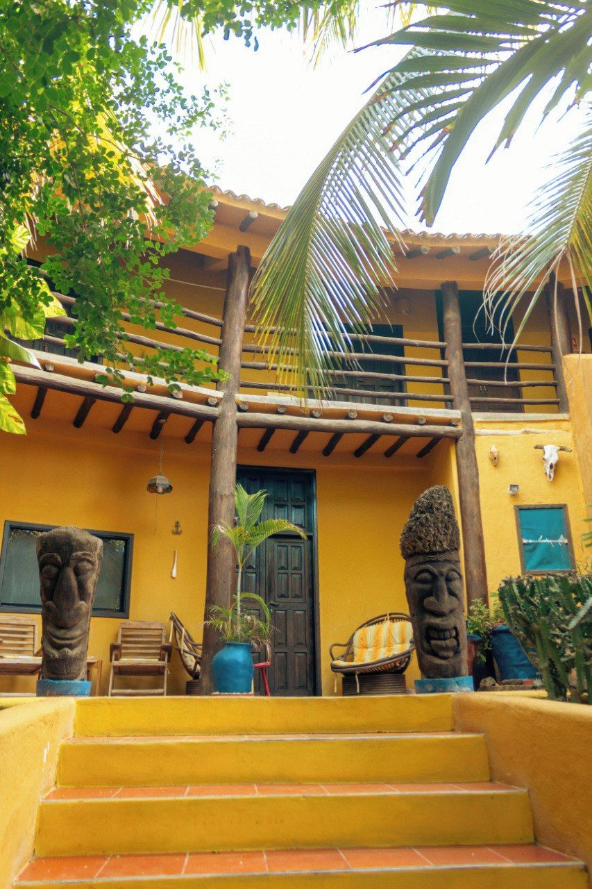 Un edificio amarillo con una escalera de madera que lleva a una puerta, dos esculturas de piedra y una palmera.