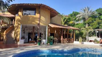 Zona de piscina con una piscina azul cristalina, un porche cubierto con asientos exteriores y una escalera que lleva a un nivel superior.