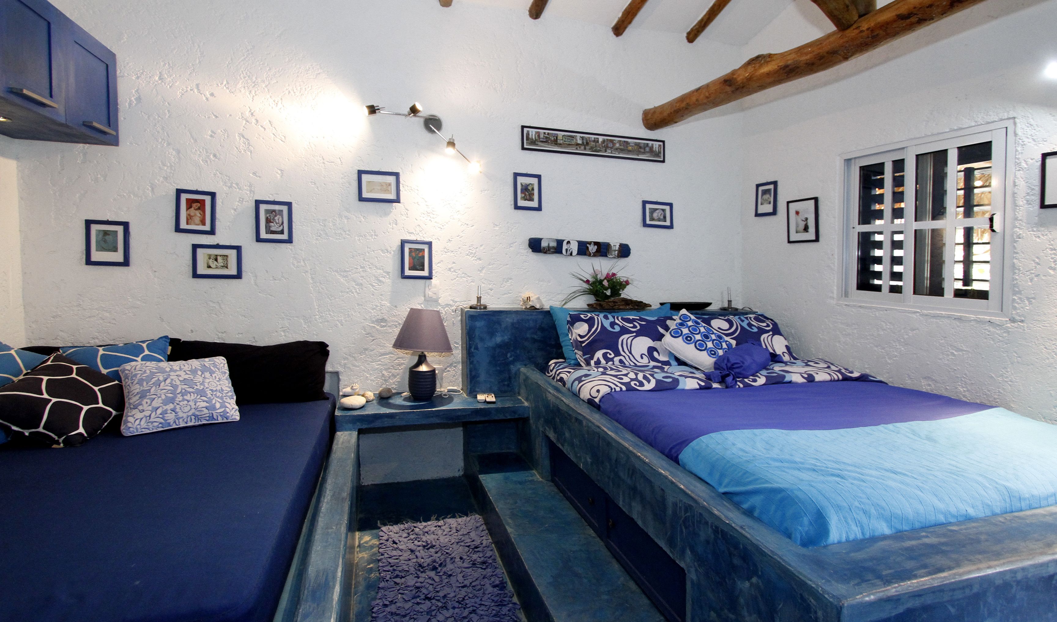 A cozy room with a bed, a sofa, and a small table with a lamp. There are framed pictures on the wall and a window with shutters.