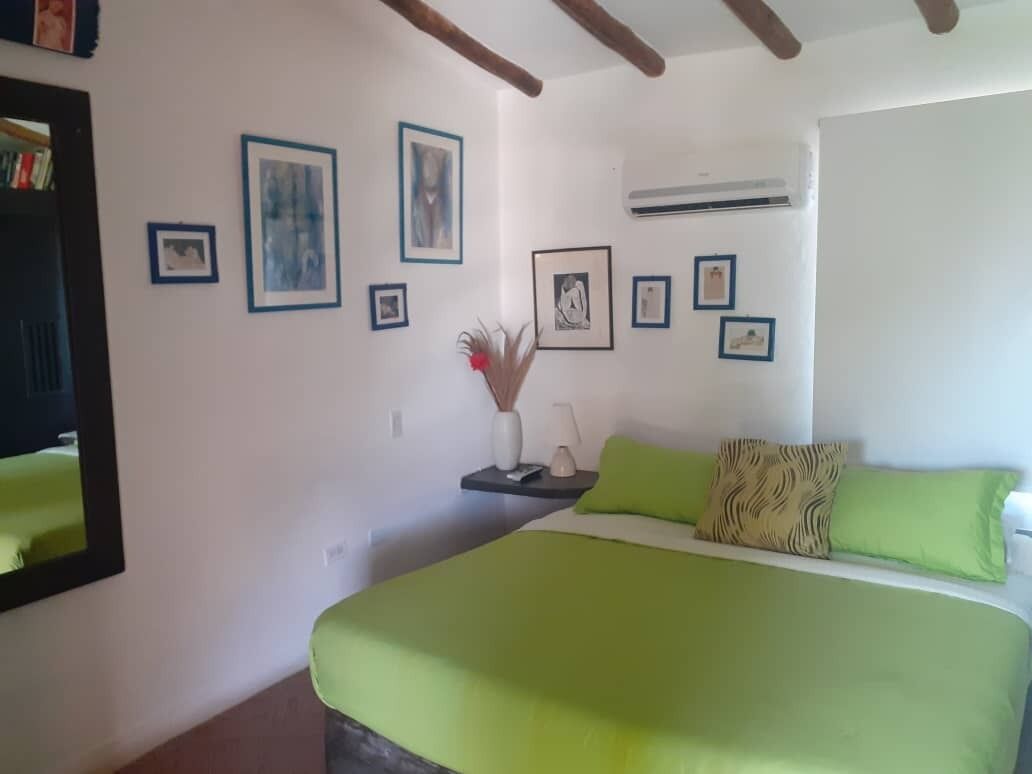 A bedroom with a bed, green bedding, a nightstand with a vase, and framed pictures on the wall.