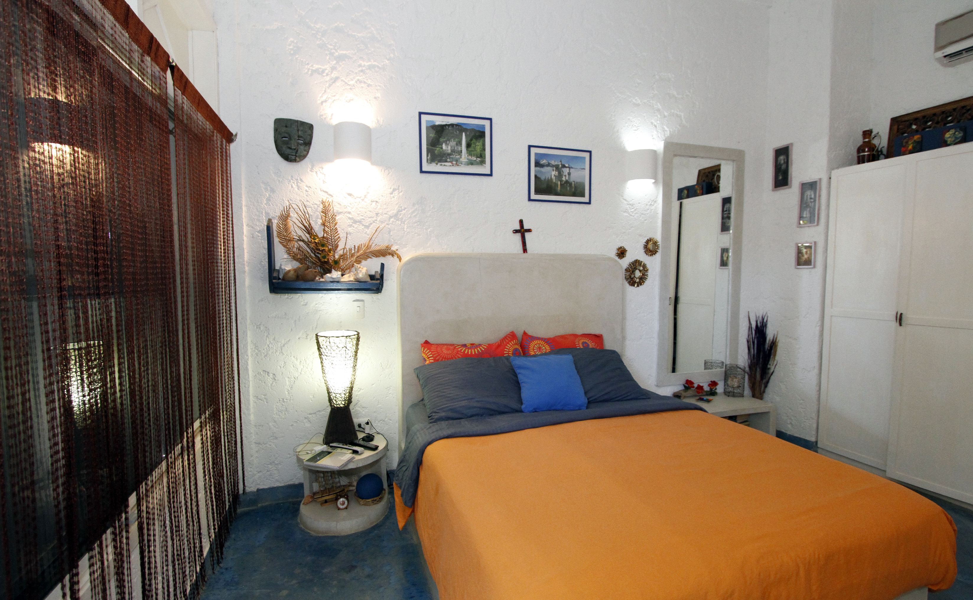 A bedroom with a bed, a nightstand, a wall-mounted shelf with decorative items, a mirror, and a door.