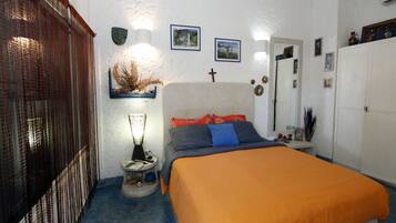 A bedroom with a bed, a nightstand, a wall-mounted shelf with decorative items, a mirror, and a door.