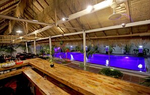 A bar area with wooden furniture, a pool with purple lighting, and a thatched roof ceiling. - Xing Angkor Hotel (Siem Reap)