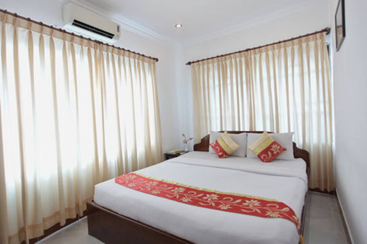 Una habitación de hotel con una cama grande, cortinas color beige y una colcha decorativa roja.