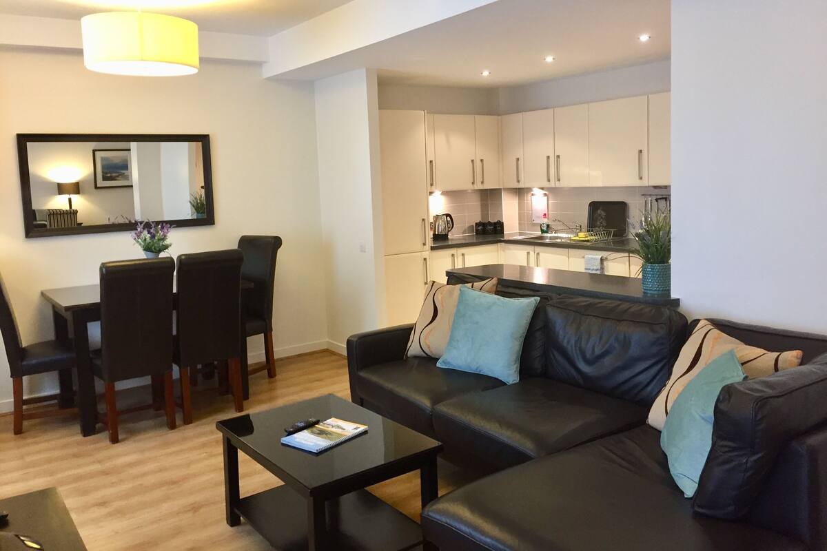 A modern living room with a black leather sofa, a coffee table, and a dining area with a table and chairs. A kitchen with white cabinets and a sink is visible in the background.