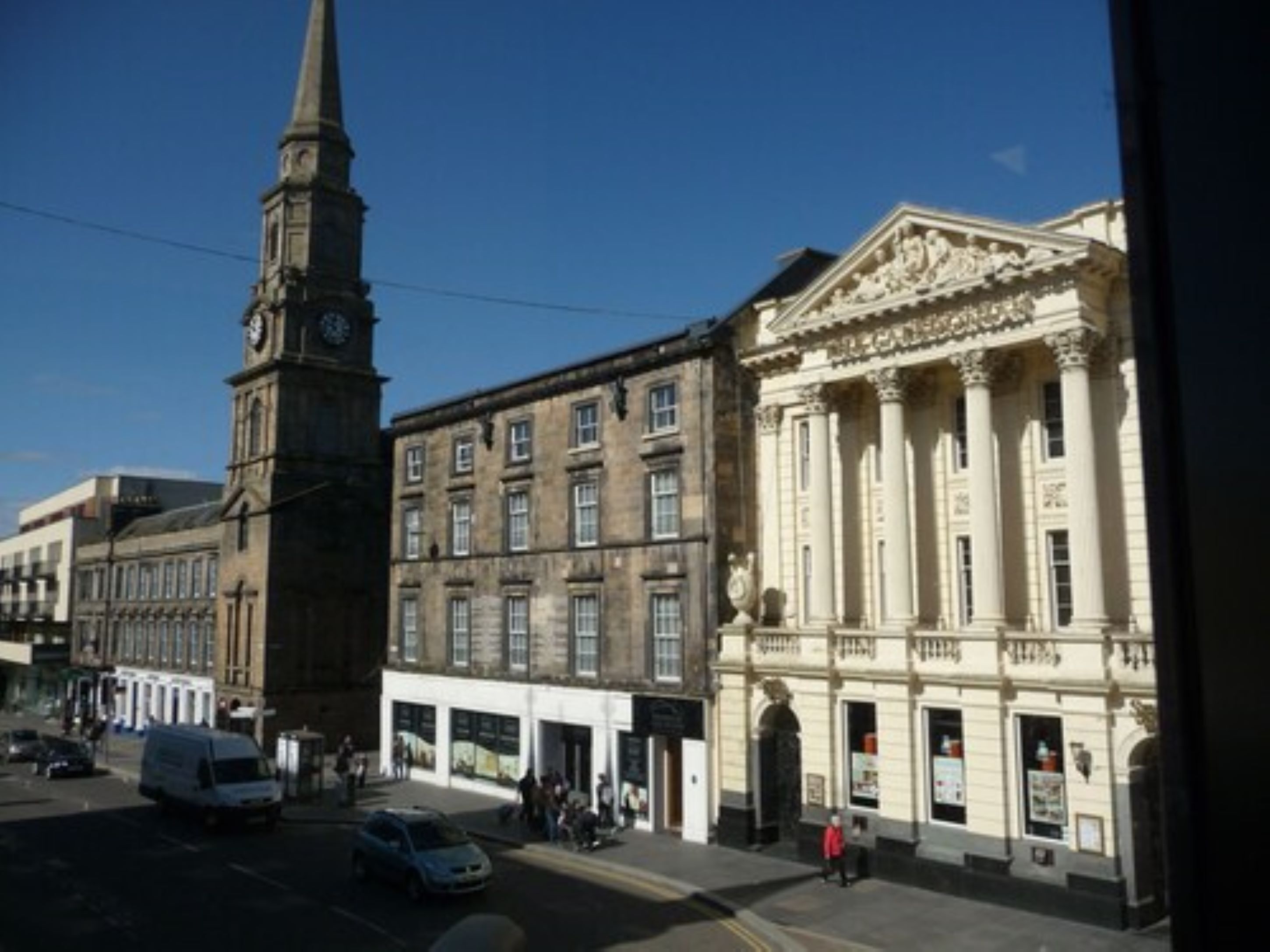 A street view with a clock tower, a building with classical architecture, and a white van parked on the side.