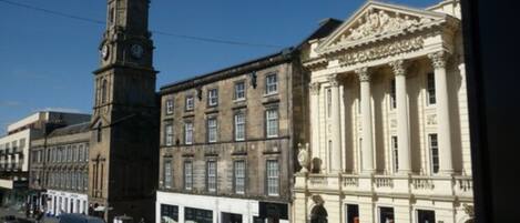 A street view with a clock tower, a building with classical architecture, and a white van parked on the side.
