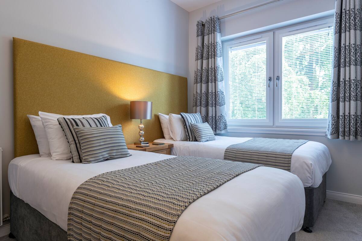 A hotel room with two beds, a headboard, bedside tables, and a window with curtains.