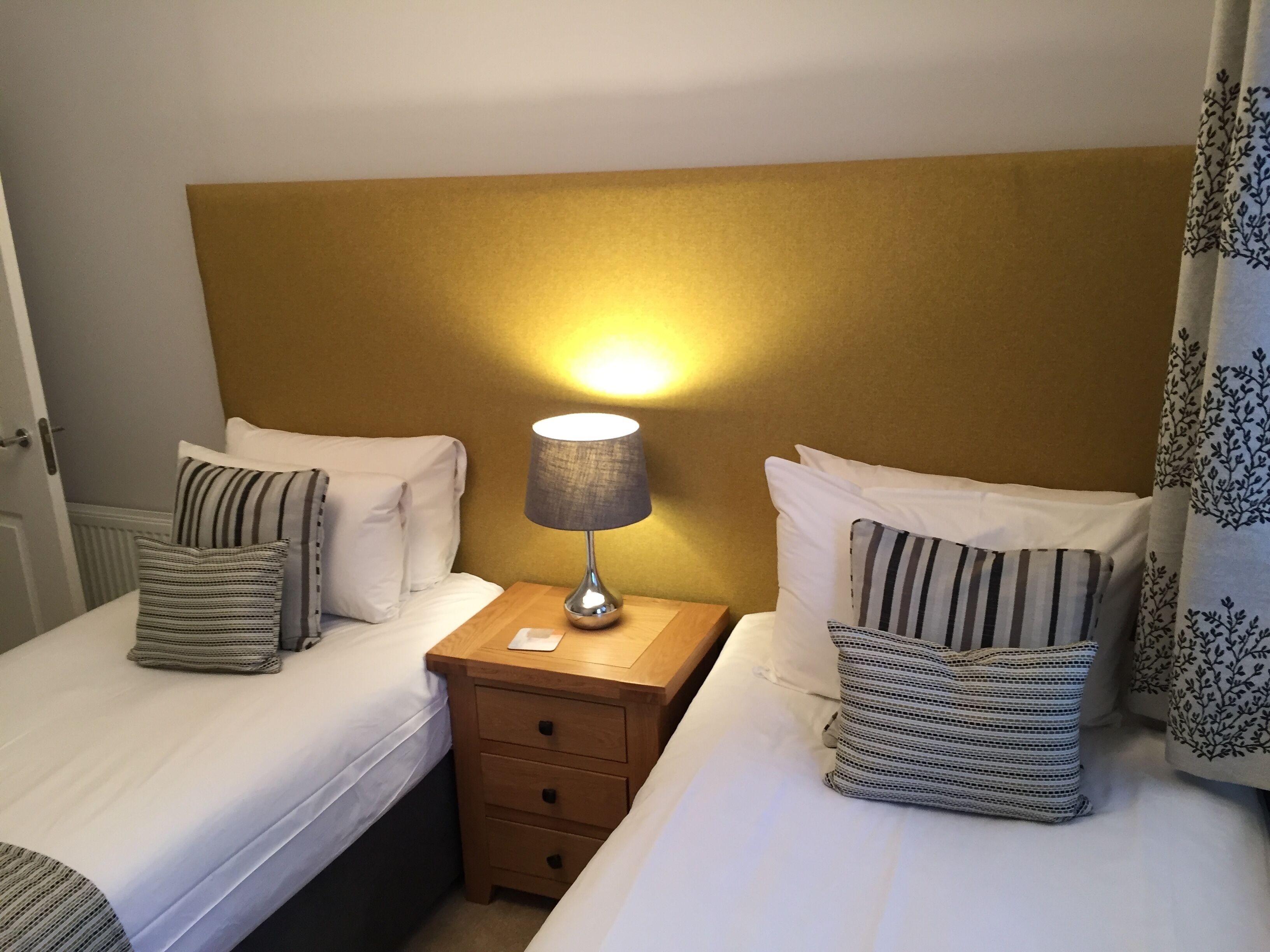A hotel room with two beds, each with striped pillows, a wooden nightstand with a lamp, and a plain wall.
