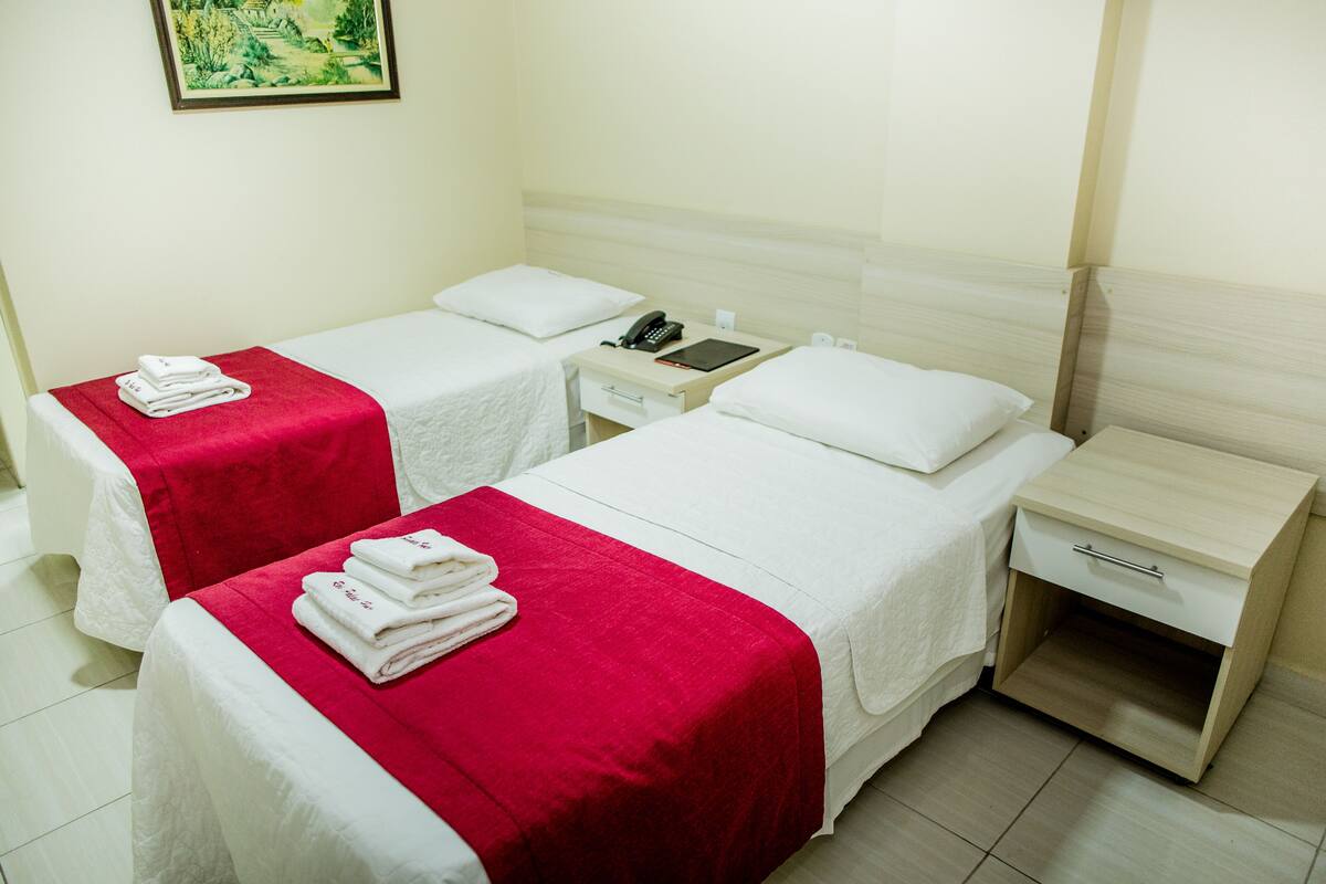 A hotel room with two single beds, each with a red and white bedspread, a bedside table with a phone and a small drawer, and a wall-mounted painting.