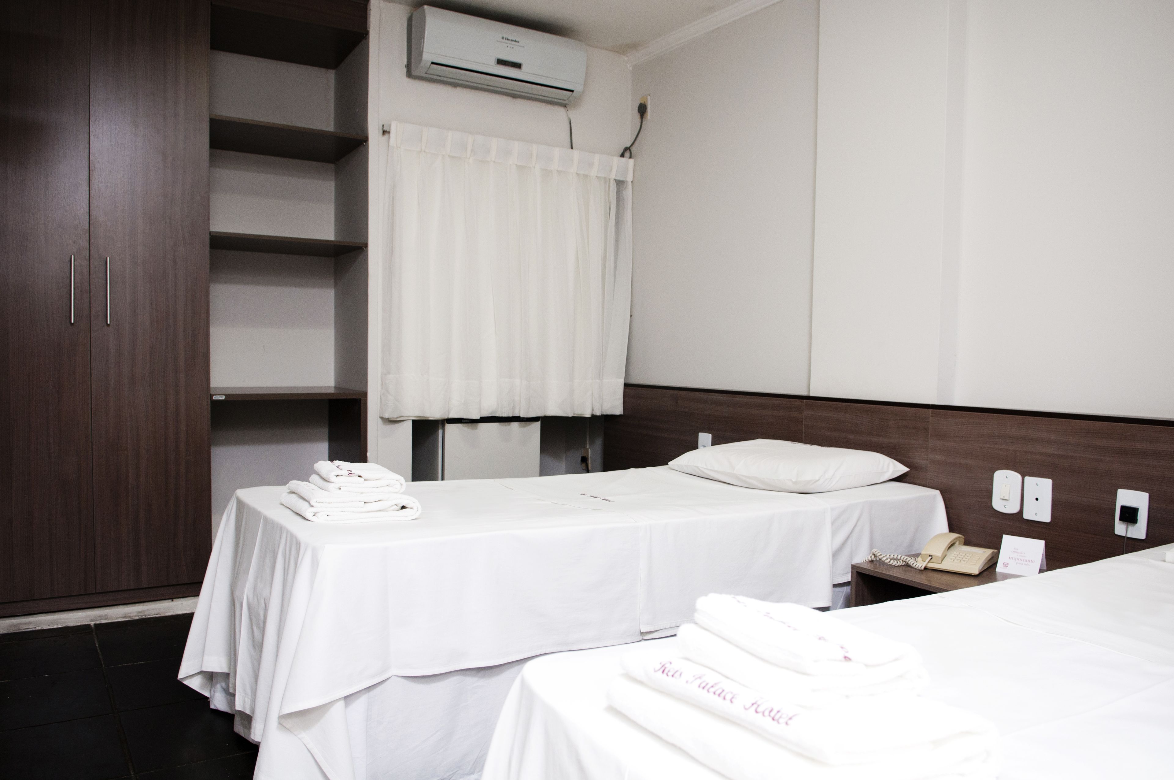 A hotel room with two beds, a wardrobe, an air conditioning unit, and a small table with a telephone.