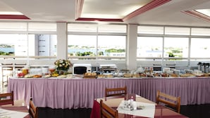 A buffet setup with a checkered tablecloth, various food items, and a view of greenery through large windows. - Reis Palace Hotel (Petrolina)