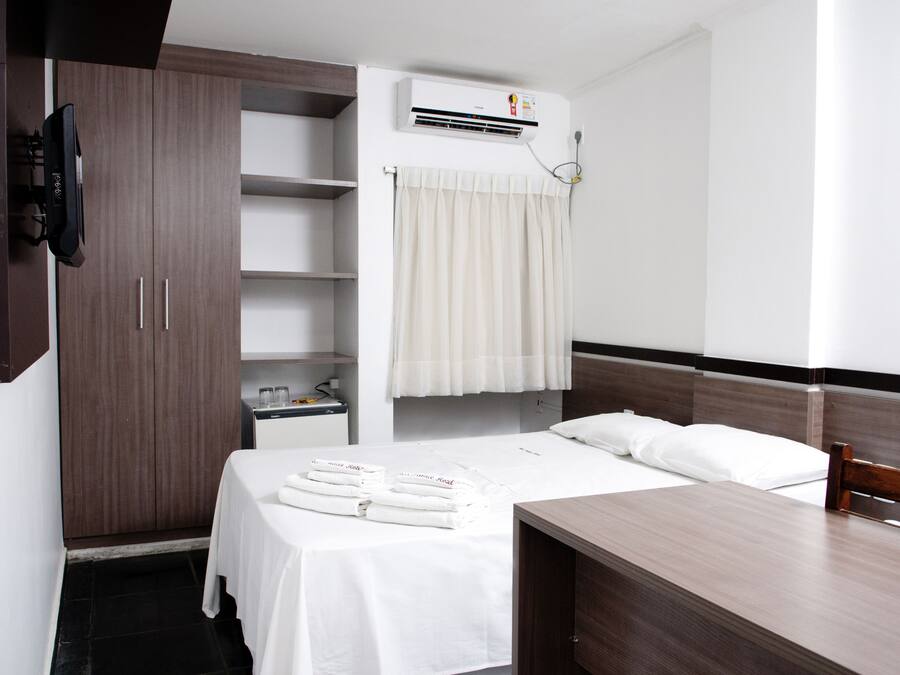 A hotel room with a bed, a desk, a wardrobe, a TV, a shelf with toiletries, and an air conditioning unit.