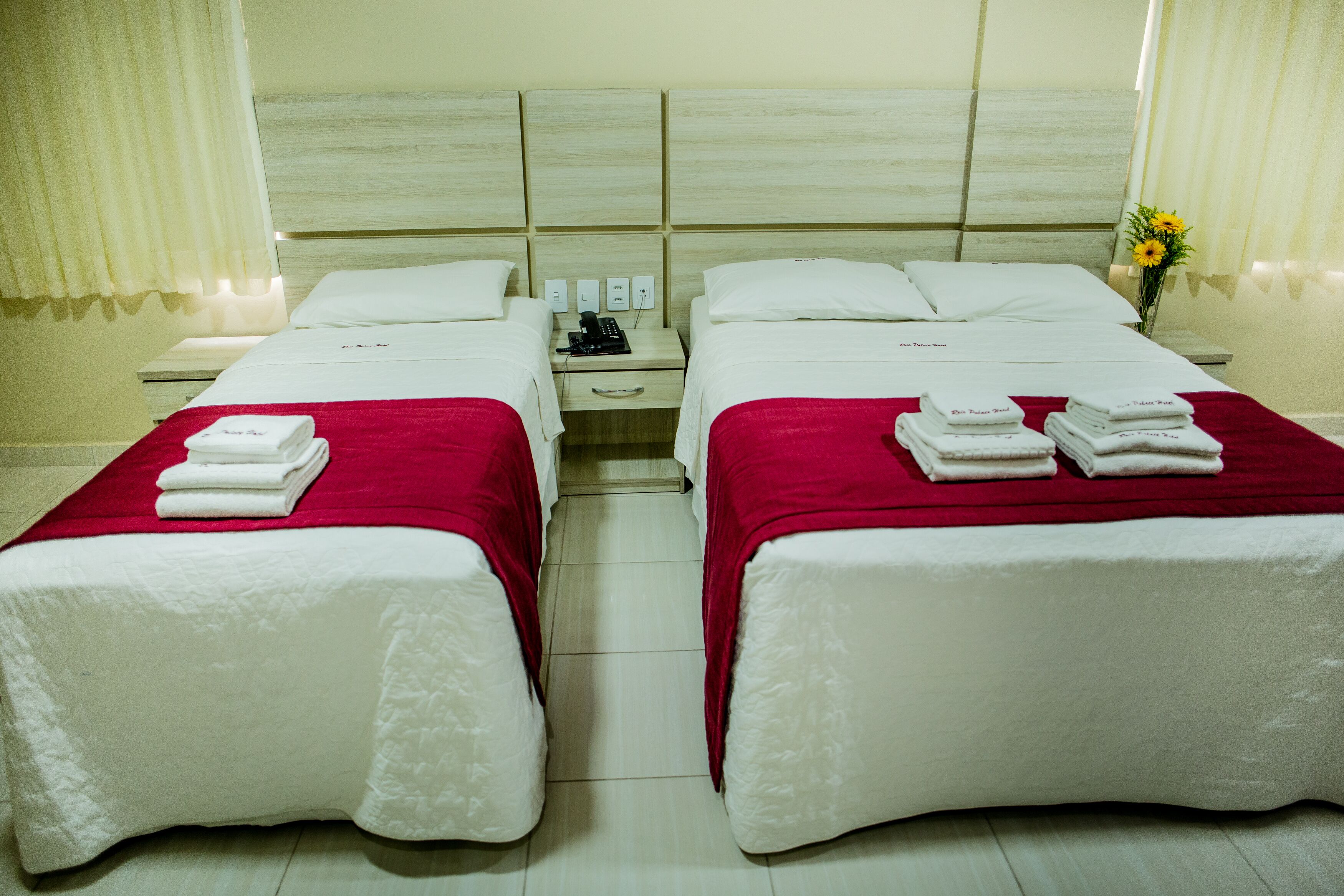 A hotel room with two beds, each with a red bedspread and white pillows. There are folded towels on each bed, and a small table with a phone and a vase with flowers.