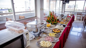 A buffet setup with various food items, a microwave, and a view of the city through large windows. - Reis Palace Hotel (Petrolina)