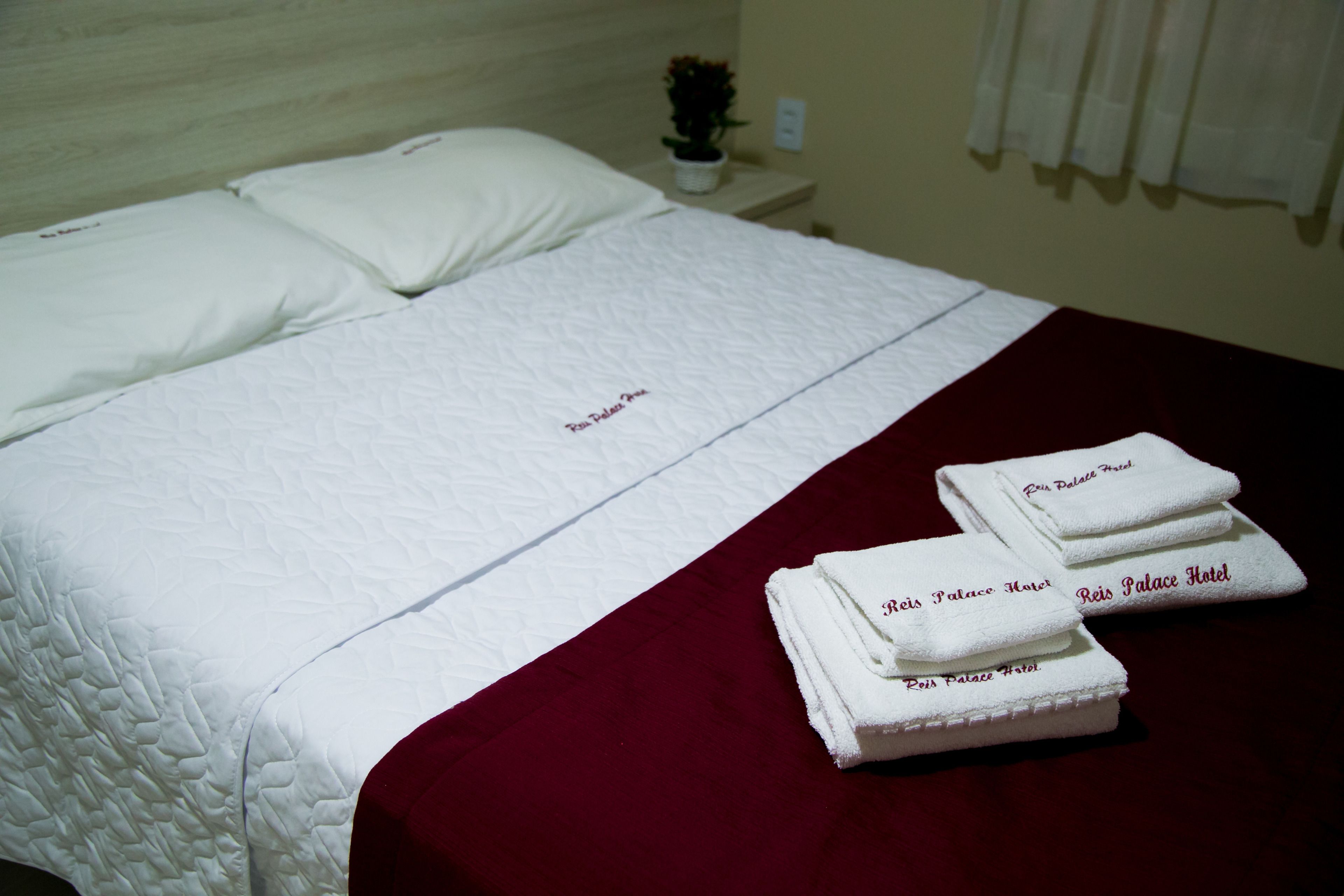 Un lit bien fait, avec du linge de lit blanc et une couverture rouge, accompagné de serviettes pliées portant le nom de l’hôtel.