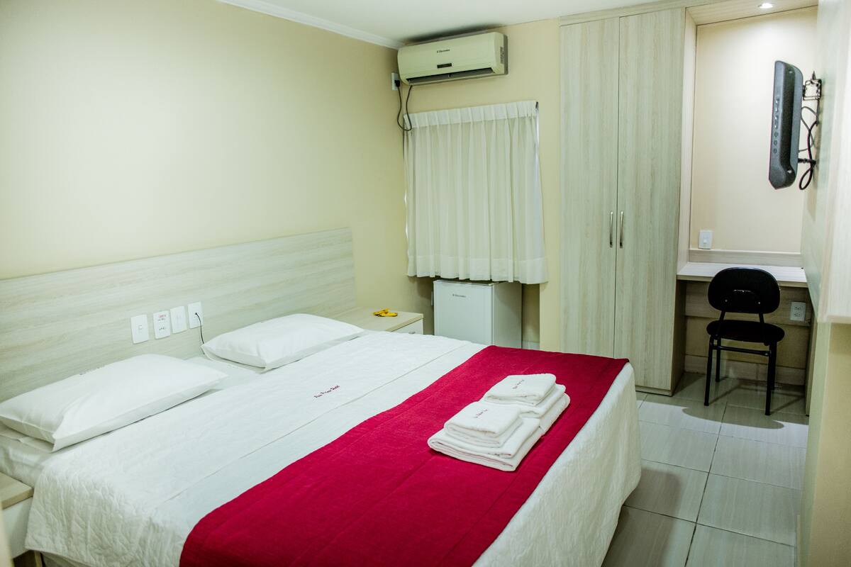 A hotel room with a bed, a desk, and an air conditioning unit.