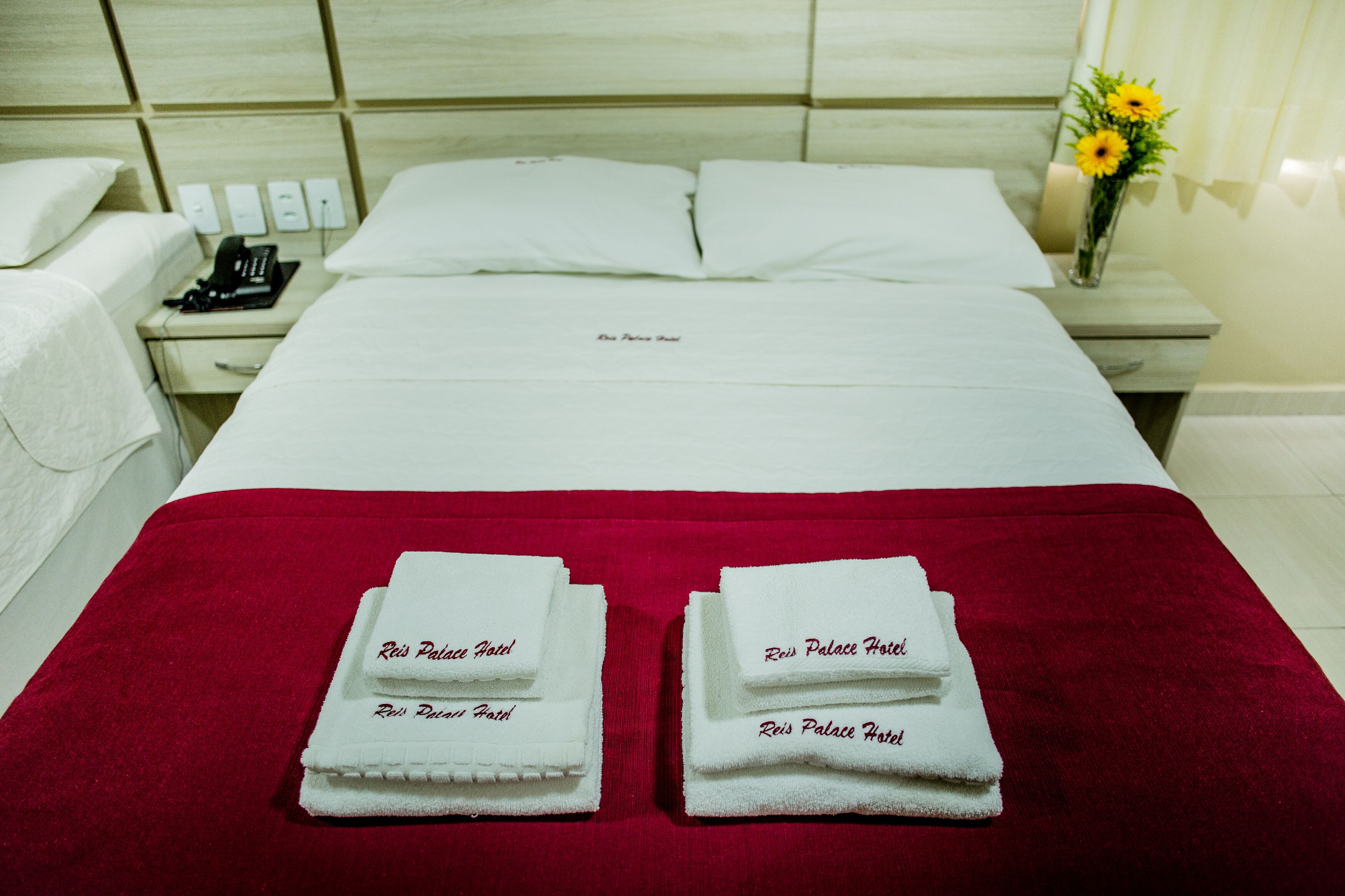 Quarto de hotel com cama bem arrumada, dois jogos de toalhas, telefone ao lado da cama e um vaso com flores.