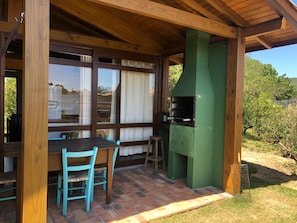 An outdoor patio with a wooden table and chairs, a green barbecue grill, and a view of the surrounding garden.
