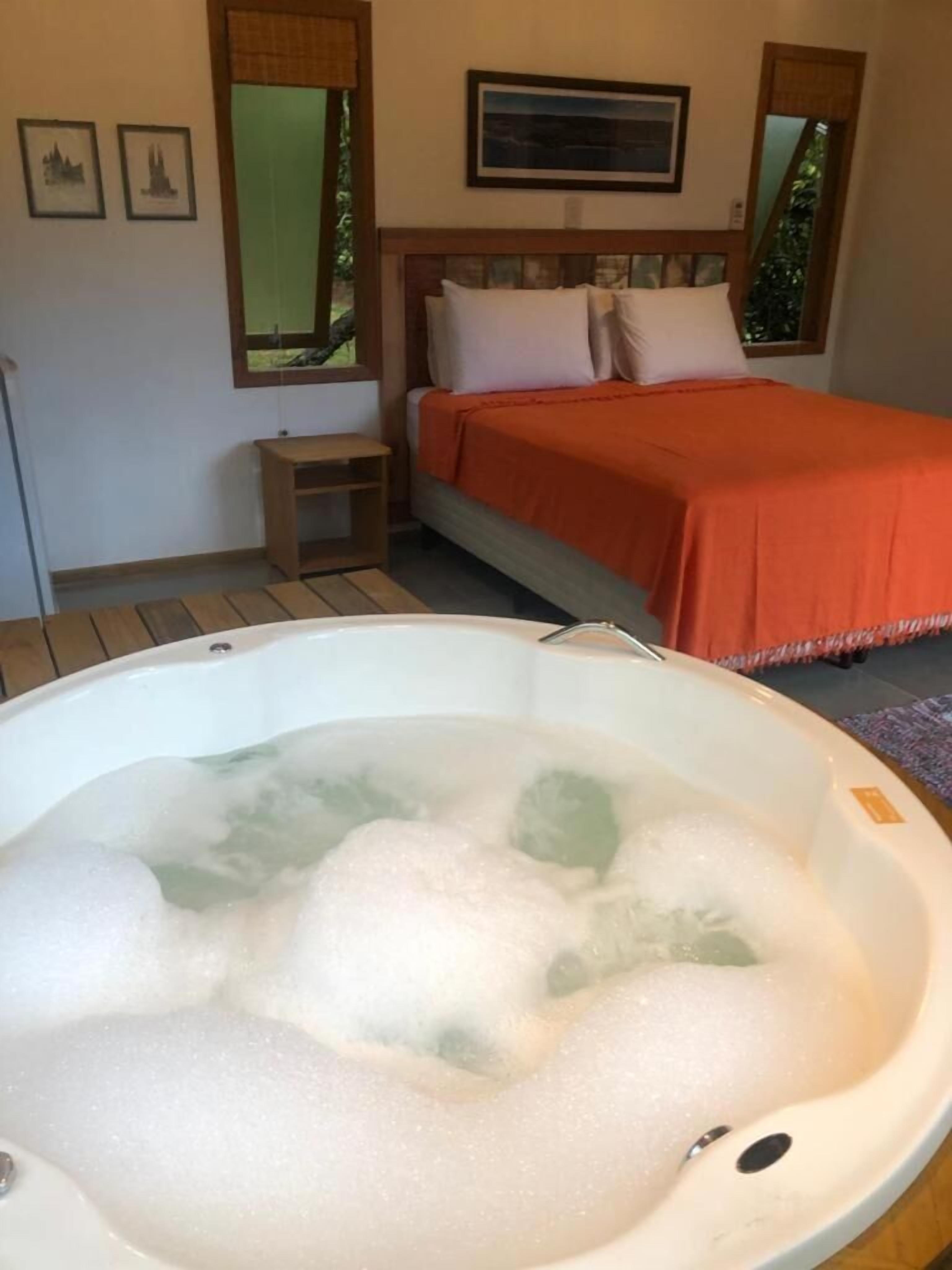 Quarto com cama, mesa de cabeceira, banheira grande cheia de espuma e janela com vista para o verde.