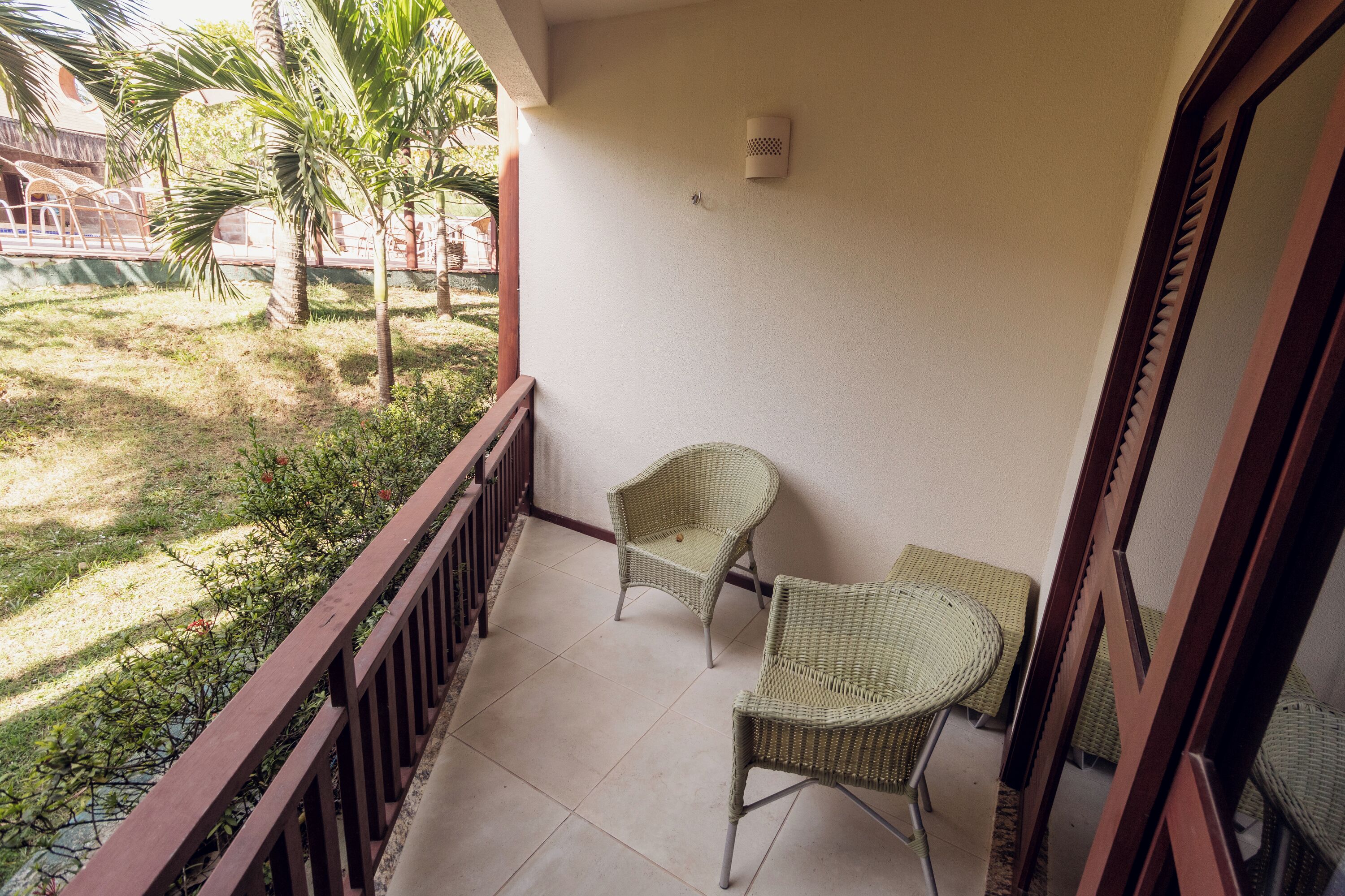Un balcon avec deux fauteuils en osier et une petite table, une balustrade en bois, et une vue sur un jardin planté de palmiers.