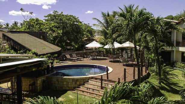 A pool area with lounge chairs and umbrellas, surrounded by palm trees and a building with a tiled roof. - Pousada Kanaloa (Jijoca de Jericoacoara)