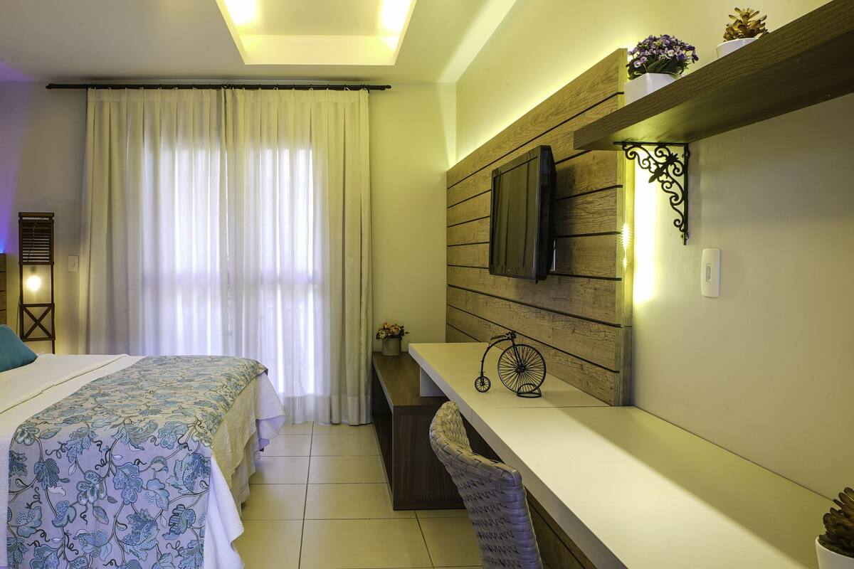 Una habitación de hotel con cama, escritorio, televisor y ventana con cortinas.