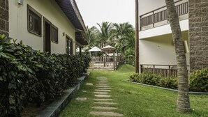 A courtyard with a stone pathway leading to a seating area with umbrellas, surrounded by greenery and buildings with balconies. - Pousada Kanaloa (Jijoca de Jericoacoara)