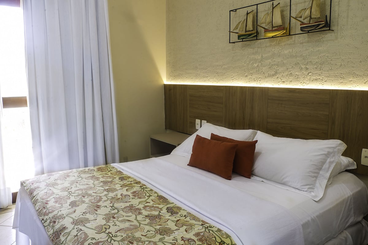 Una habitación de hotel con cama, cortinas blancas, cabecero de madera y una obra de arte colgada en la pared.