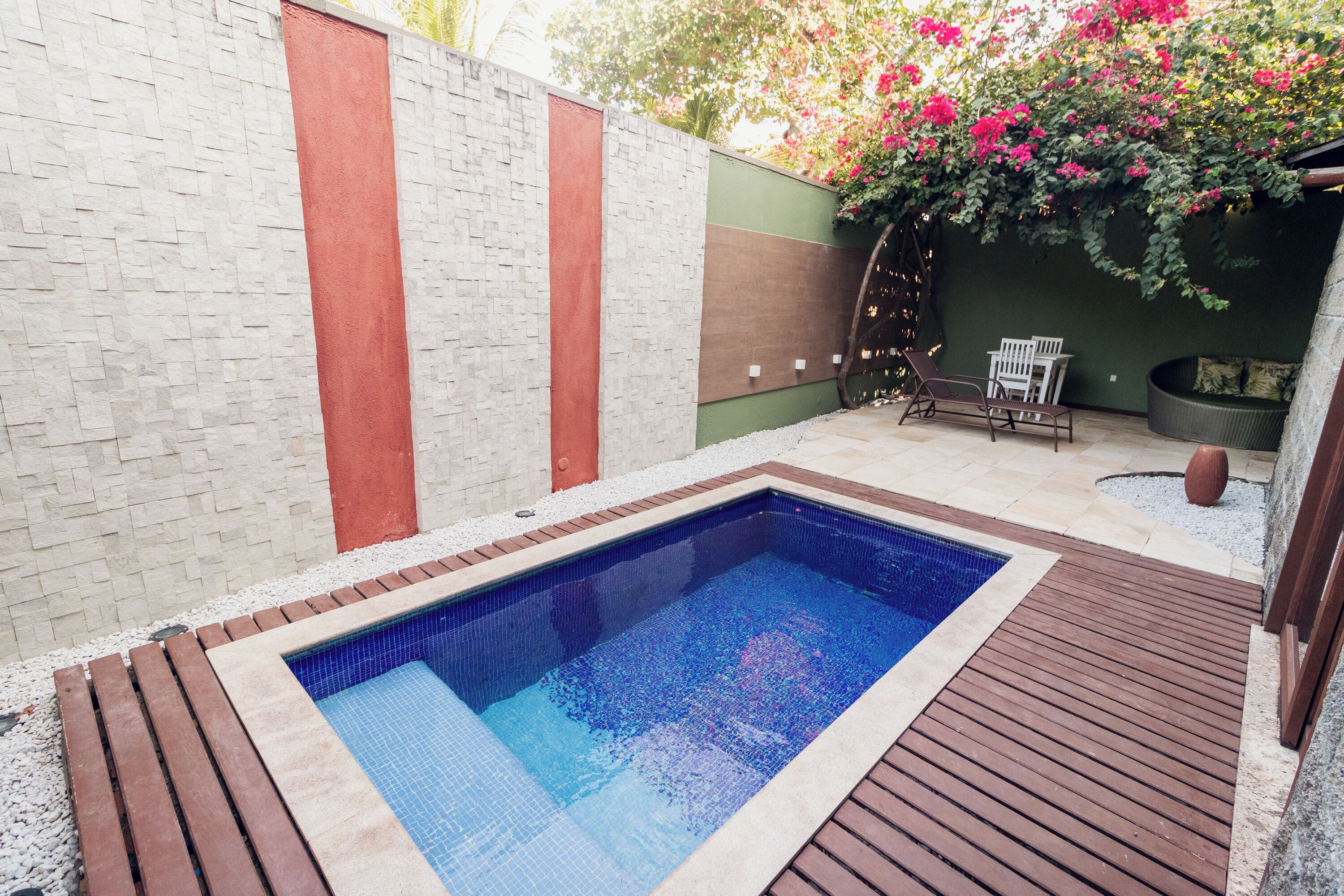 A rectangular swimming pool with a wooden deck surrounding it, a white chair, and a green wall with a climbing plant.