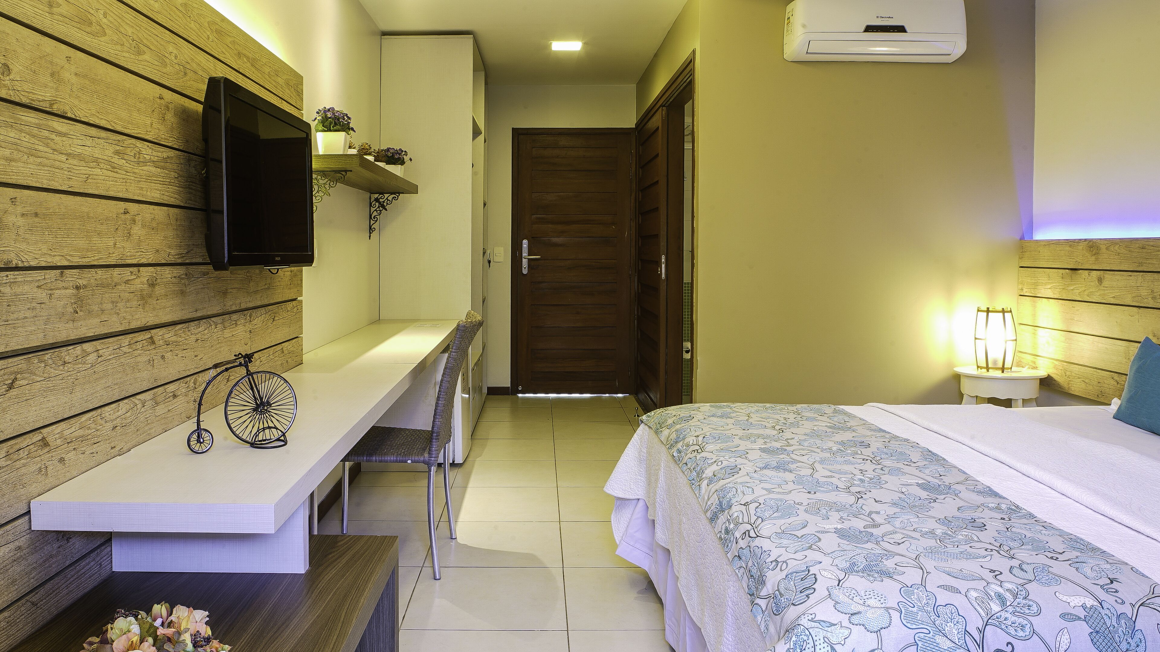 A bedroom with a bed, bedside table, chair, desk, TV, and an air conditioning unit.