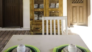 A rustic wooden table with two white plates and a white chair in front. Behind the table, there is a wooden cabinet with glass doors displaying various dishes. A wooden door and a bulletin board with pictures are also visible. - Pousada Kanaloa (Jijoca de Jericoacoara)