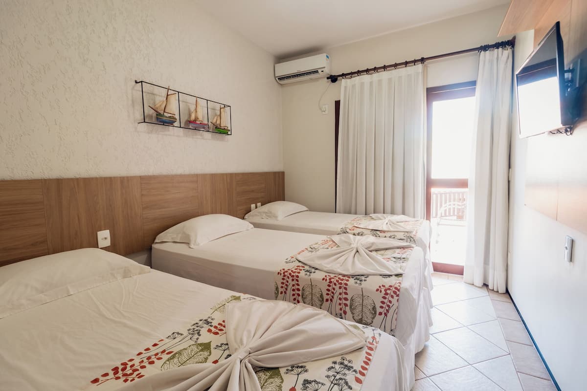 Habitación de hotel con tres camas, cabecera de madera, aire acondicionado, televisor y balcón con cortinas.