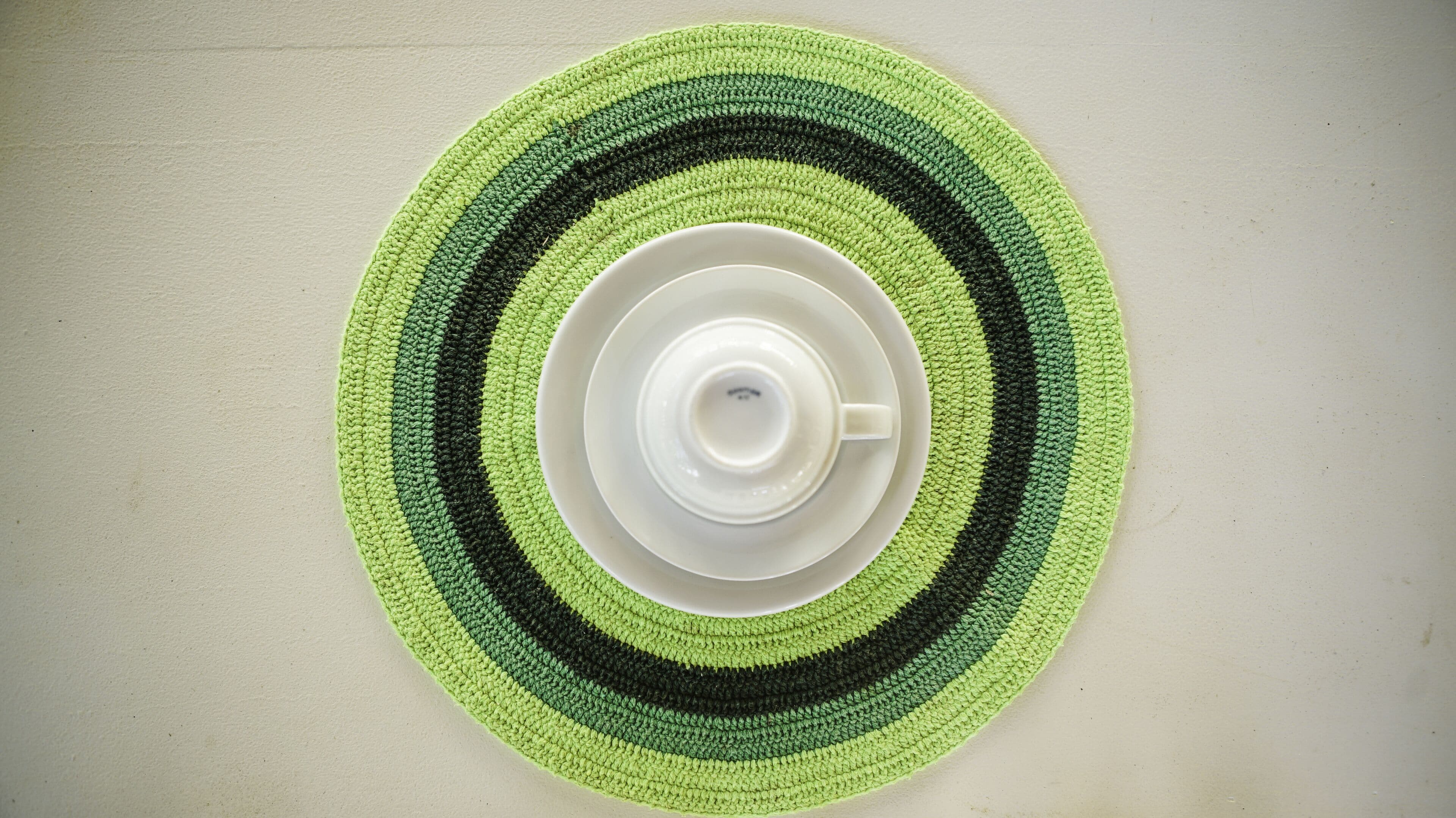 A white cup and saucer centered on a green and black striped placemat on a plain wall.