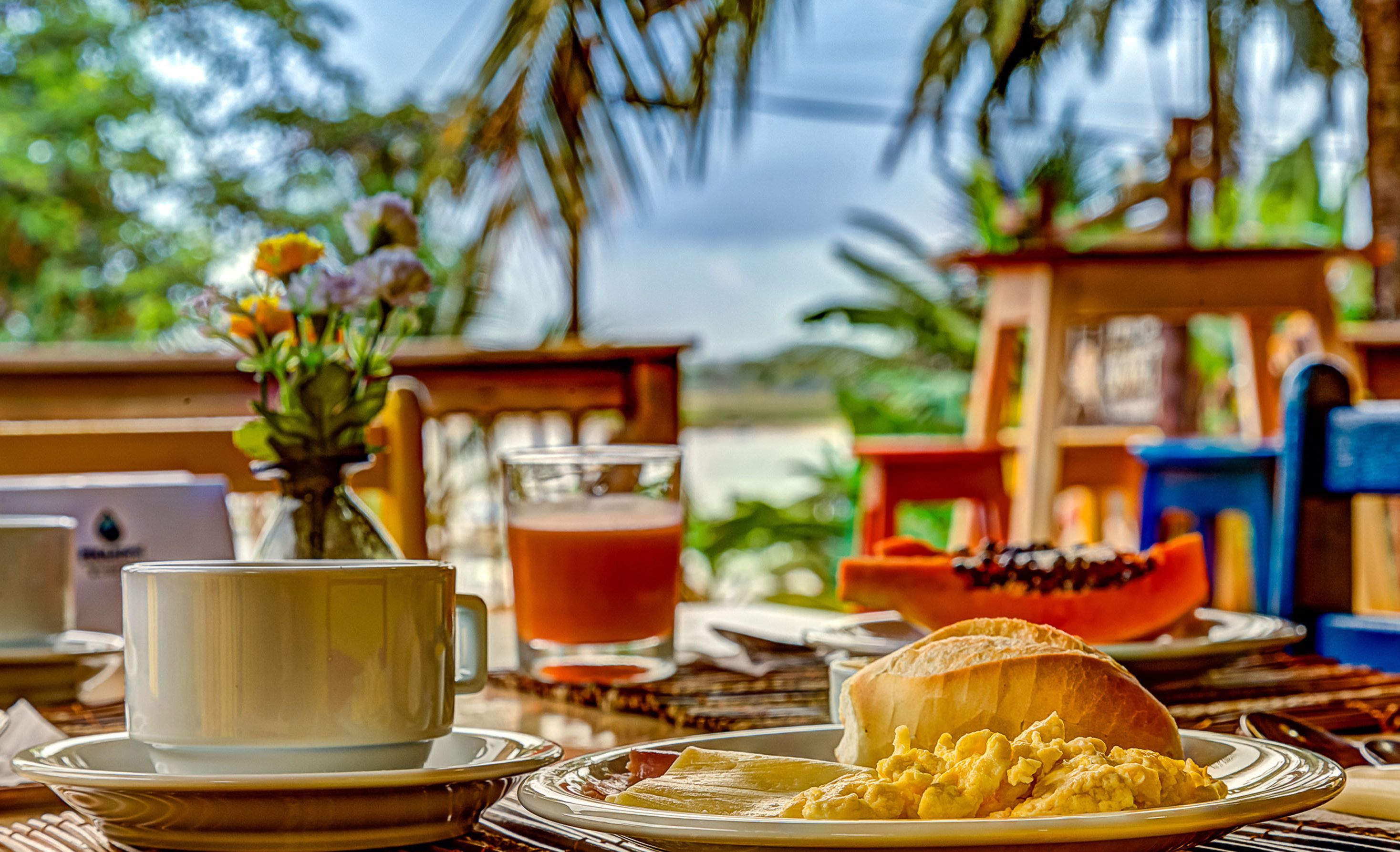 Colazione con una tazza, un bicchiere di succo, uova strapazzate e un panino su un tavolo con uno sfondo tropicale.