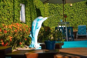 A dolphin statue in a pool area with a lush green hedge backdrop and a small table with chairs.