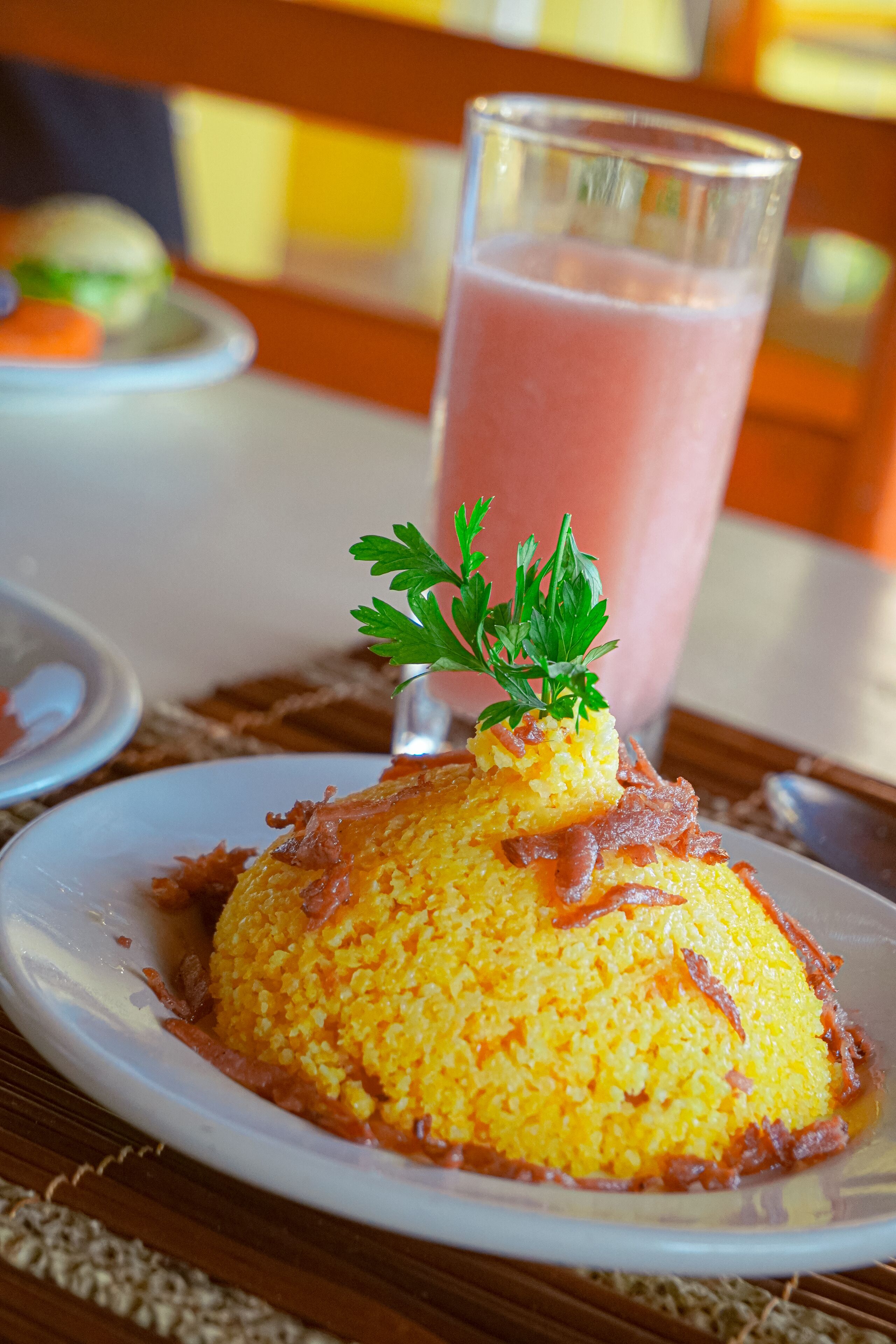 A plate of yellow rice topped with bacon and parsley, with a glass of pink beverage in the background.