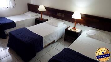 Quarto de hotel com três camas individuais, cada uma com colchão branco e cobre azul. Há duas mesas de cabeceira com luminárias e o chão é revestido de azulejos.