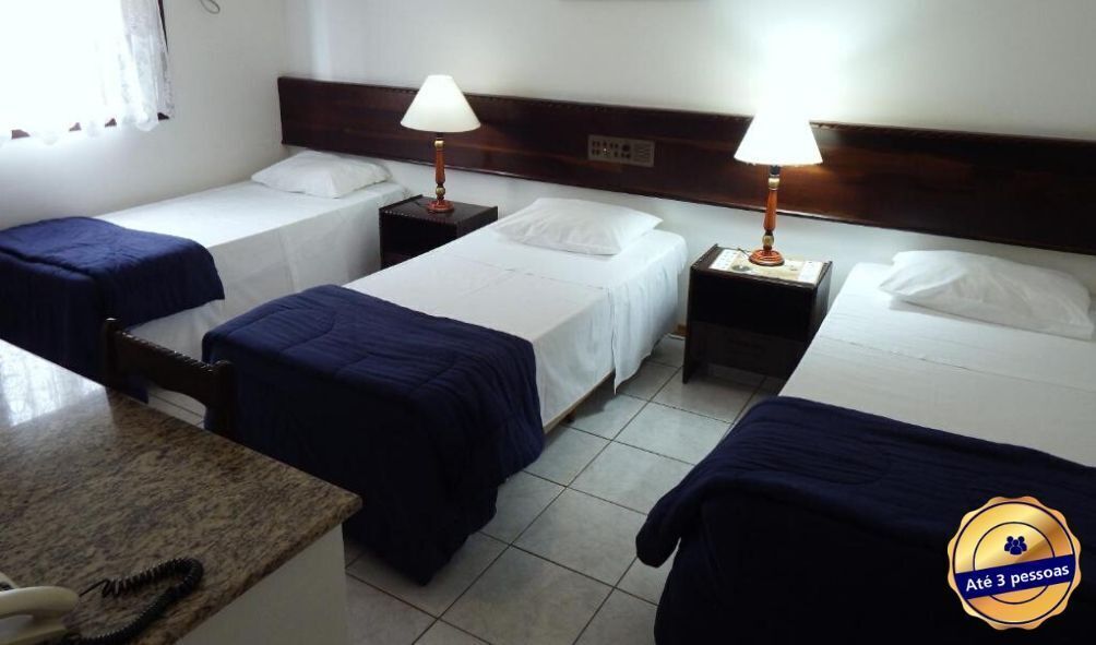 A hotel room with three single beds, each with a wooden headboard and white bedding. There are two bedside tables with lamps, and a small desk with a telephone.