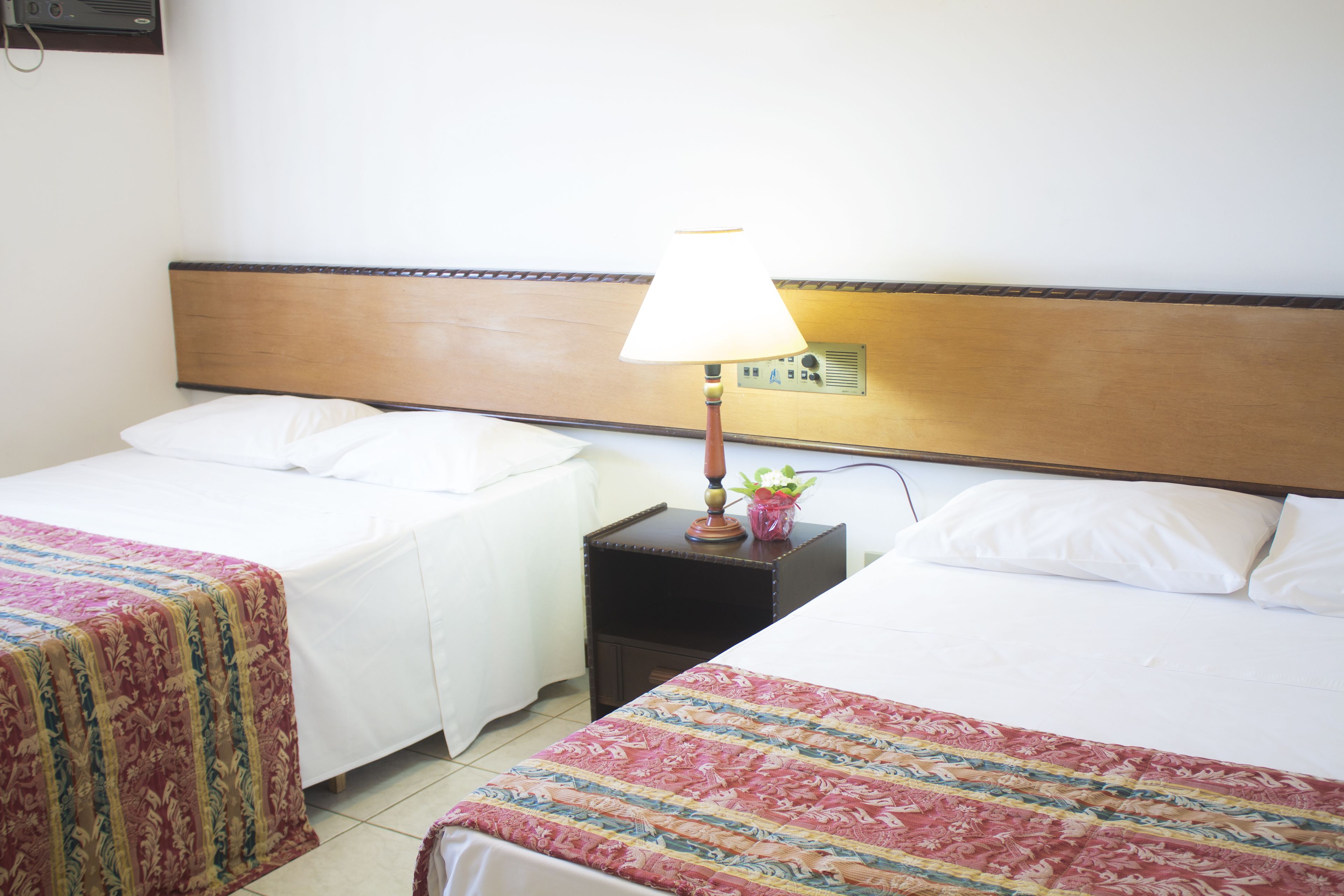 A hotel room with two single beds, a nightstand with a lamp, and a wall-mounted control panel.