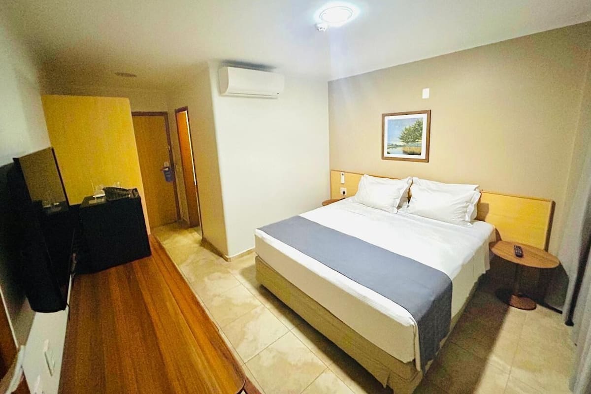 A hotel room with a bed, bedside table, and a small table with a chair. There is a TV on a stand and a door leading to another room.