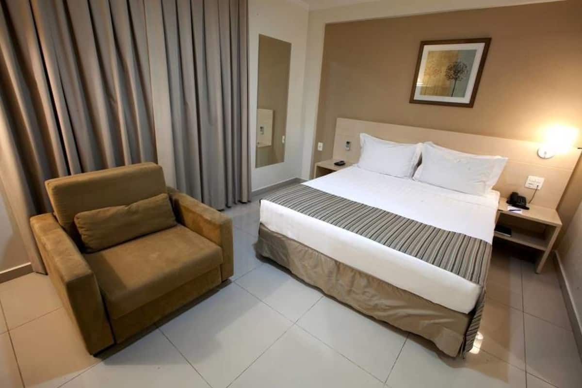 A hotel room with a bed, a chair, a nightstand, and a wall-mounted lamp.