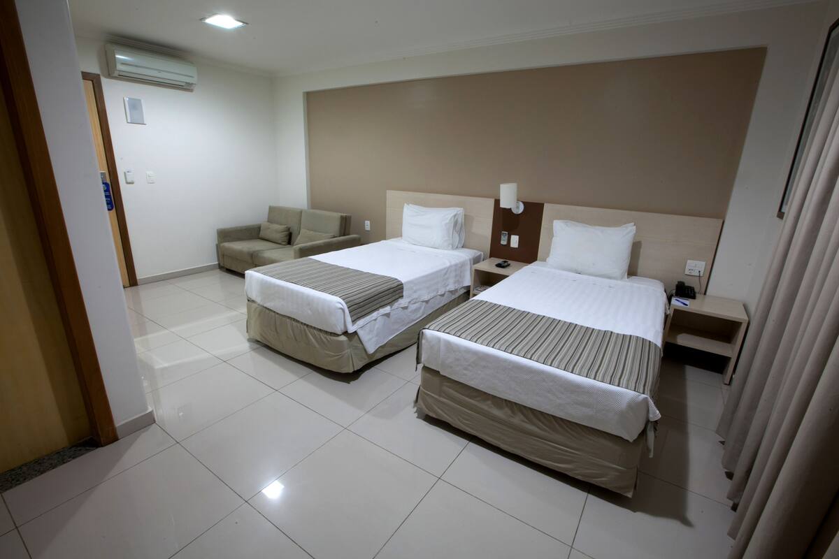 A hotel room with two beds, a sofa, a nightstand, and an air conditioning unit.