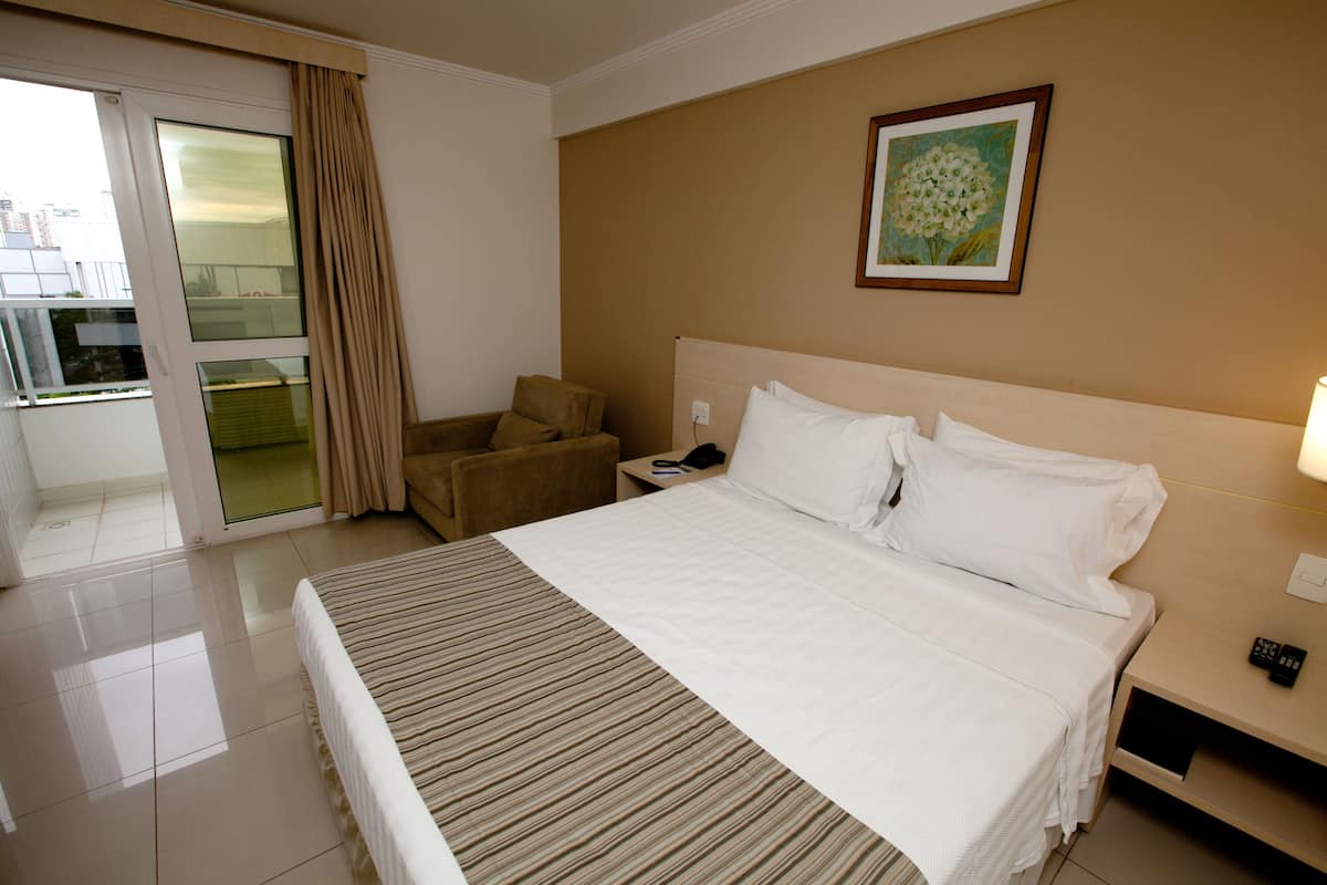 A hotel room with a large bed, a chair, a nightstand, a lamp, and a painting on the wall.