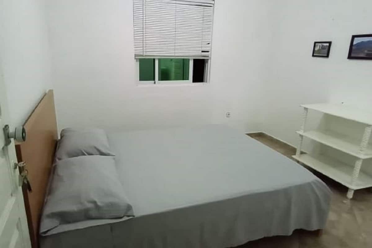 A small, single-bed room with a wooden headboard, a window with blinds, a white shelf unit, and a few framed pictures on the wall.