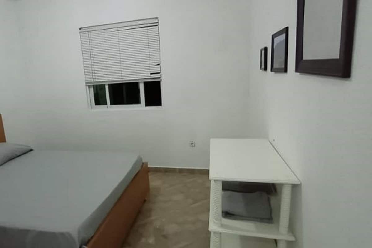 A bedroom with a bed, a window with blinds, a small white nightstand, and a wall-mounted picture frame.