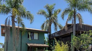 A two-story house with a green exterior, a balcony, and a fenced yard with palm trees. - Pousada Vale Verde Bungalows (Imbituba)